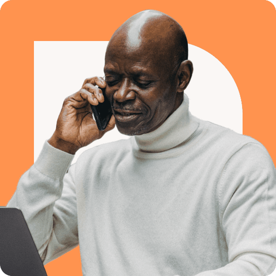Ein älterer schwarzer Mann in einem weißen Pullover spricht mit einem Smartphone, vor dem ein Laptop aufgeklappt ist, vor einem orangefarbenen Hintergrund.